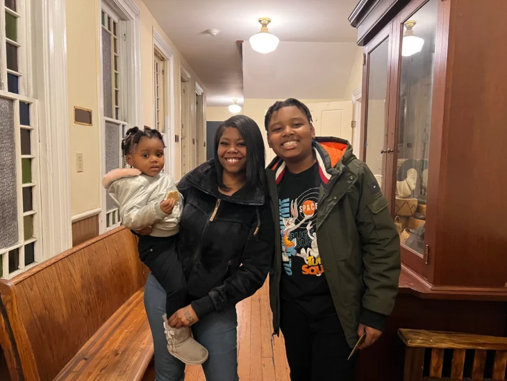 an african american family of a mother with her two children are smiling at the camera they are in an office and are wearing casual winter clothes