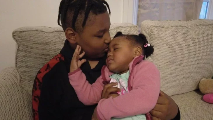 a brother kisses his little sister that he is carrying in his arms the boy is sitting in a cream armchair and both are wearing casual winter clothes