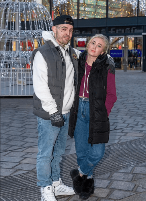 una pareja posndo en una plaza donde se ve un árbol de Navidad de fierro cubierto de luces la pareja viste de manera informal