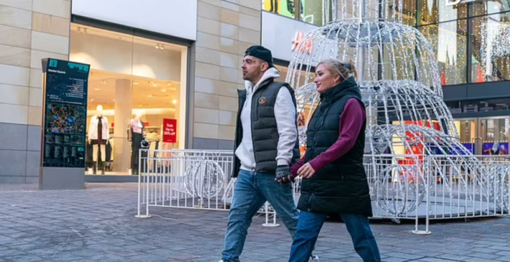 una pareja caminando en una plaza van tomados de la mano llevan ropa deportiva invernal casual se observa una tienda departamental al fondo de la imagen