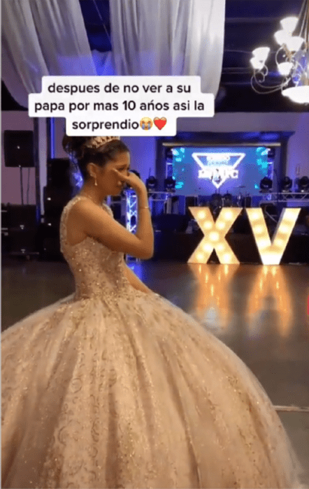 una quinceañera llora en su fiesta de XV años lleva un vestido estilo princesa de color crema largo con tul y lleva una tiara sobre la cabeza acomodada con un moño al fondo se ven encendidas las letras XV 