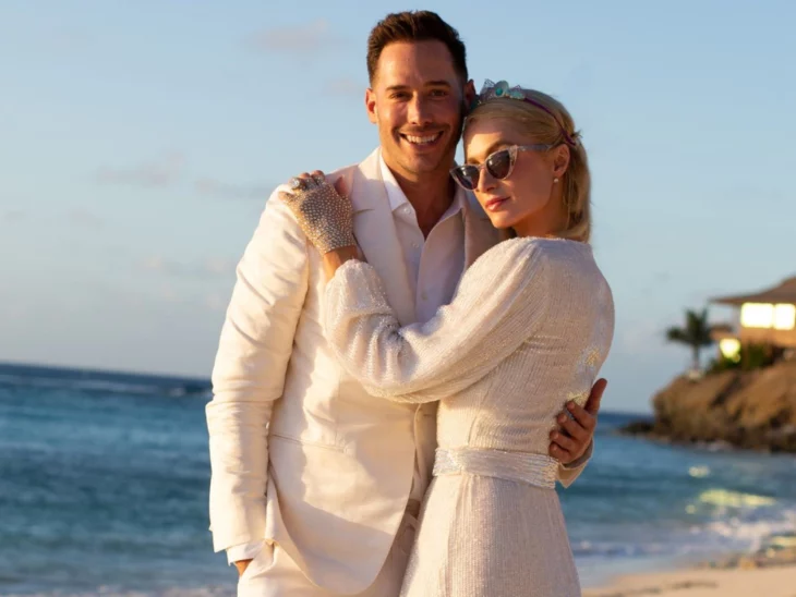 Paris Hilton y su esposo Carter Reum posando a la orilla de la playa abrazados y vestidos de blanco ella trae lentes de sol y guantes brillosos el un traje delgado con camisa blanca sin corbata 
