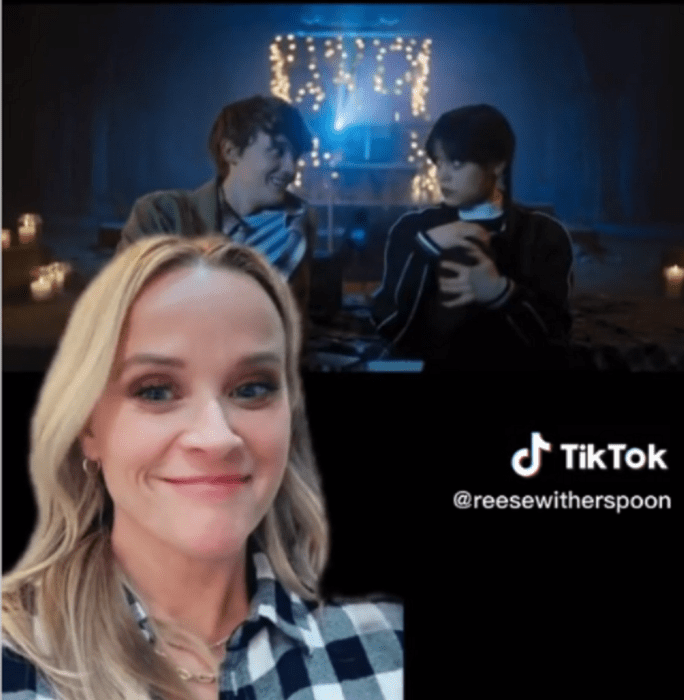 image from TikTok where Reese Witherspoon reacts to the Merlina scene where they mention her movie Legally Blonde, Reese wears a black and white plaid shirt, long blonde hair loose behind her, the characters of Merlina and Tyler appear