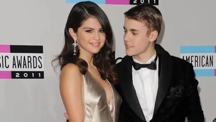 Selena Gomez y Justin Bieber cuando eran pareja posando en una alfombra roja ella lleva un vestido de noche de color crema y el cabello suelto y peinado en ondas su maquillaje es discreto él lleva un esmoquin negro con una camisa blanca 