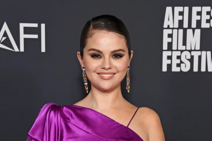 The singer Selena Gomez posing on a red carpet is wearing a fuchsia evening dress, her hair tied up and very long golden earrings, her makeup is evening