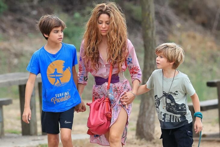 imagen de la cantante Shakira junto a sus hijos Milán y Sasha caminado tomados de la mano por un parque ella lleva un vestido floreado y una bolsa roja los chicos visten con ropa casual playeras y shorts 