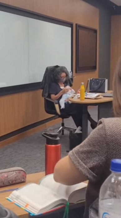 una profesora alimenta con un biberón a un bebé mientras está sentada frente a su escritorio durante un examen que realiza a sus alumnos lleva ropa de color oscuro 