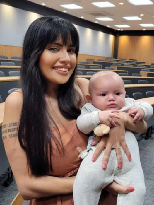 una madre joven sostiene a su bebé en brazos están dentro de un aula de la universidad ella lleva un vestido color arena sin mangas y el bebé un mameluco gris 
