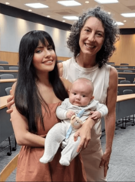 imagen dentro del aula de una Universidad dos mujeres posan en la foto una de ellas sostiene a un bebé en brazos los tres llevan ropa casual