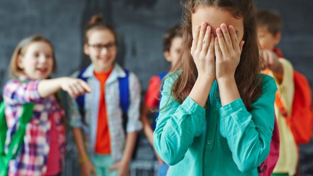 Niña con blusa azul tapándose los ojos y niños señalándola