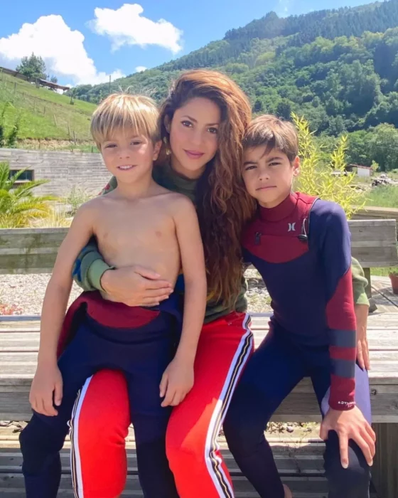 Shakira junto a sus hijos Shasa y Milan sentados sobre una banca de madera con trajes para surfear la cantante lleva ropa deportiva en color rojo y verde 