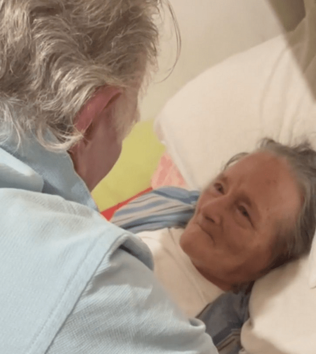 dos abuelitos en una habitación, la señora está acostada en la cama viendo al abuelo que se acerca a ella