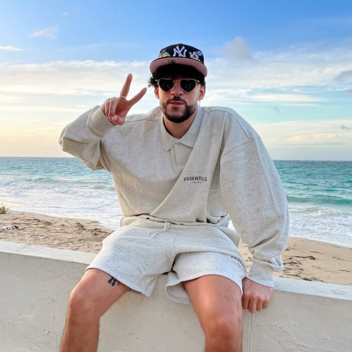 Fotografía de Bad Bunny sentado en el malecón de una playa saludando con la señal de amor y paz 