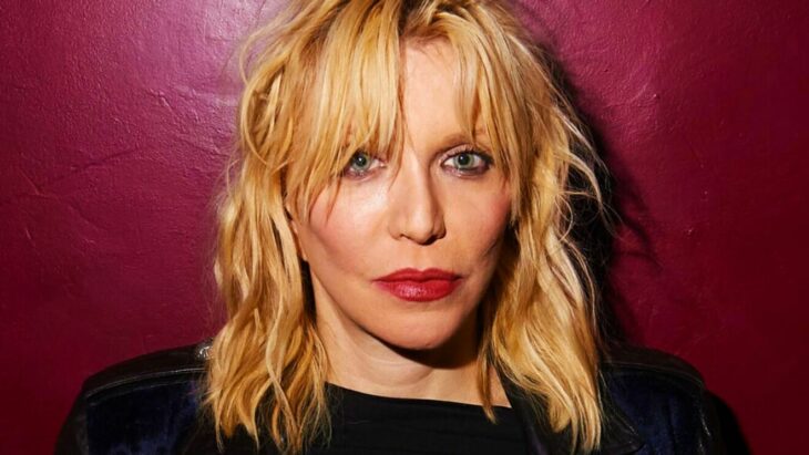 Courtney Love en primer plano lleva el cabello desaliñado rubio medio corto y un maquillaje muy marcado trae una blusa negra