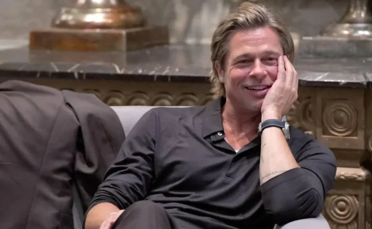 el actor Brad Pitt está sentado en un sillón en una rueda de prensa lleva ropa informal de tono oscuro está sonriendo y sostiene su rostro con una mano