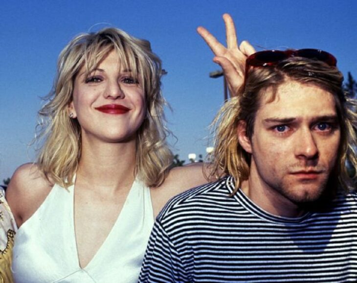 Courtney Love y Kurt Cobain en una alfombra roja el músico luce algo serio mientras que su esposa sonrie y hace la señal de la victoria ella lleva un vestido blanco de satén y el una sudadera de rayas