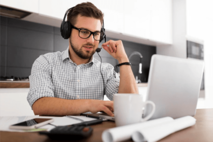 un joven con una diadema para hablar por teléfono con manos libres frente a una computadora tiene a un lado una taza de café blanca