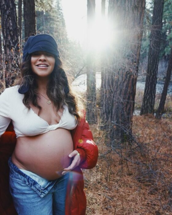 actriz Gina Rodríguez-LoCicero posando con su barriga de embarazo en un bosque rodeada de árboles 