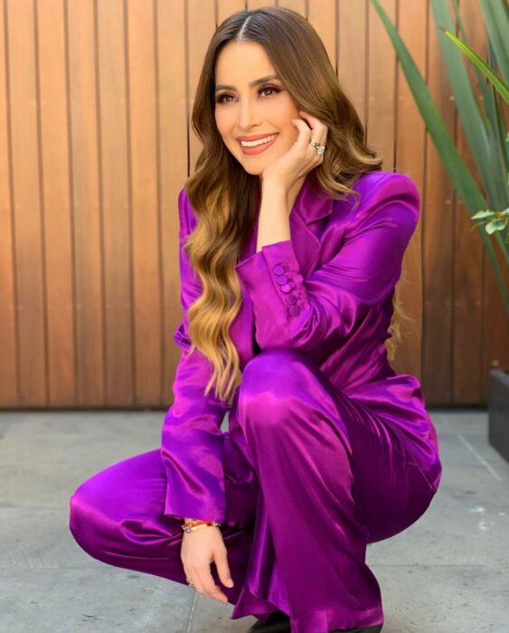 Cynthia Rodríguez posando en cuclillas vestida con un conjunto de satín en color morado 