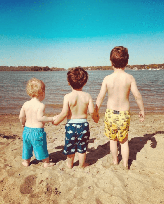 tres pequeños niños tomados de la mano y viendo al mar llevan shorts