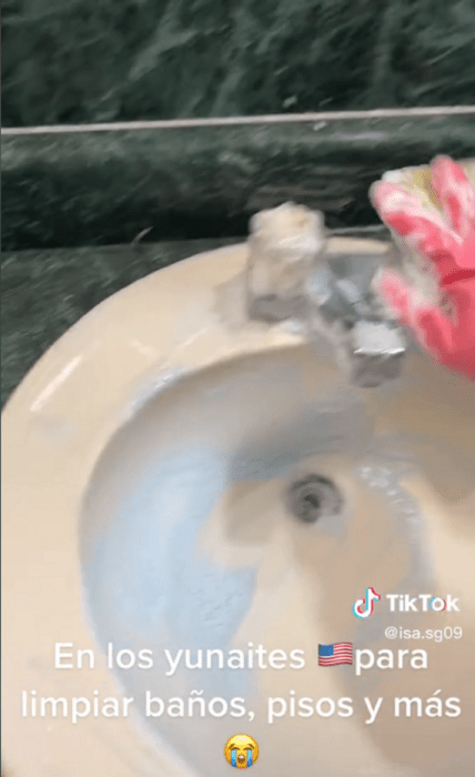 imagen donde se aprecia que una persona con guantes rosas está lavando con jabón un lavabo trae una esponja en la mano con la que lo talla se ve parte del azulejo en color verde y el lavabo es blanco