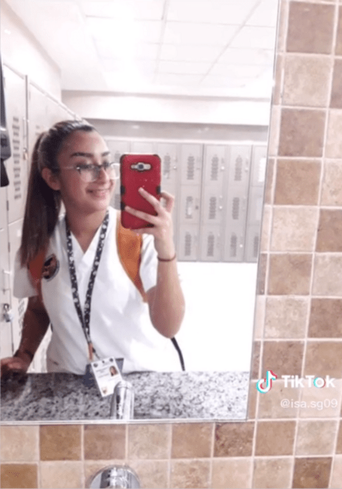 un chica se toma una foto frente al espejo lleva un traje de enfermera y una credencial colgada al cuello está soenrieno y lleva el cabllo recogido en una coleta su celular tiene una funda roja