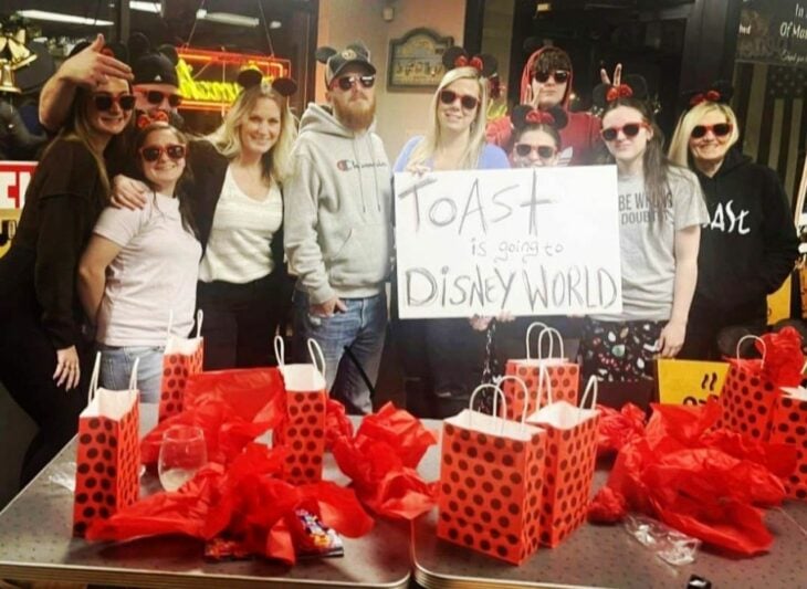 empleados de una cafetería sostienen una pancarta con un anuncio en ingles todos visten de manera informal se aprecia también una mesa con diferentes bolsas de regalo de color rojo sobre una mesa 