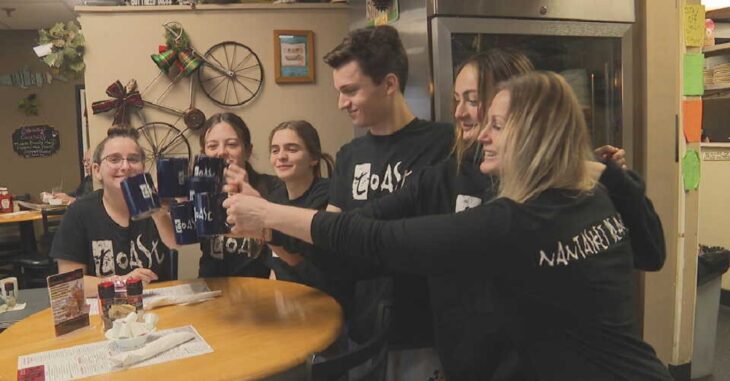 Employees in black uniforms toast with navy blue coffee cups in a restaurant They are seated around a round table in the back You can see a bicycle as a wall decoration and a large refrigerator