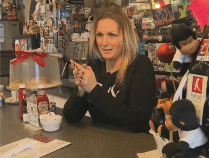 una mujer esta de pie atrás del mostrador de una cafetería lleva un uniforme de sudadera negra con letras blancas al frente es rubia tiene el cabello largo y lacio lleva un maquillaje discreto