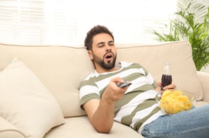 un hombre tirado en un sofá mientras bosteza lleva un recipiente con botanas y un refresco de cola en la mano y con la otra mano está cambiando canales con el control de la TV
