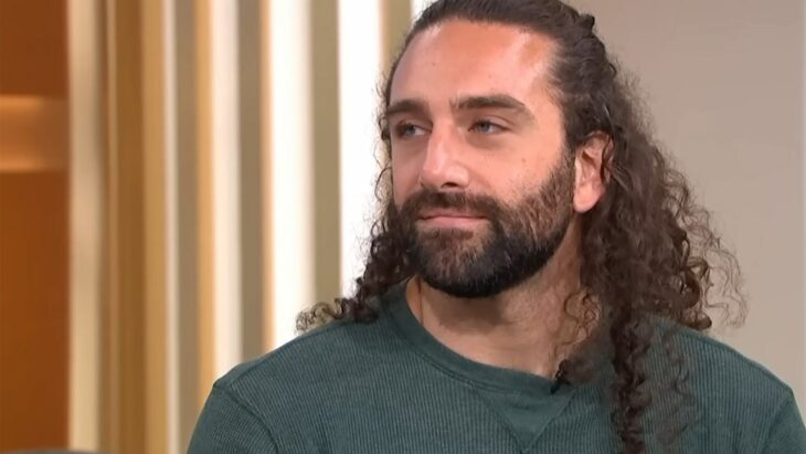 a man with long brown hair with a beard and a mustache is wearing an olive green sweatshirt and looking away from the camera