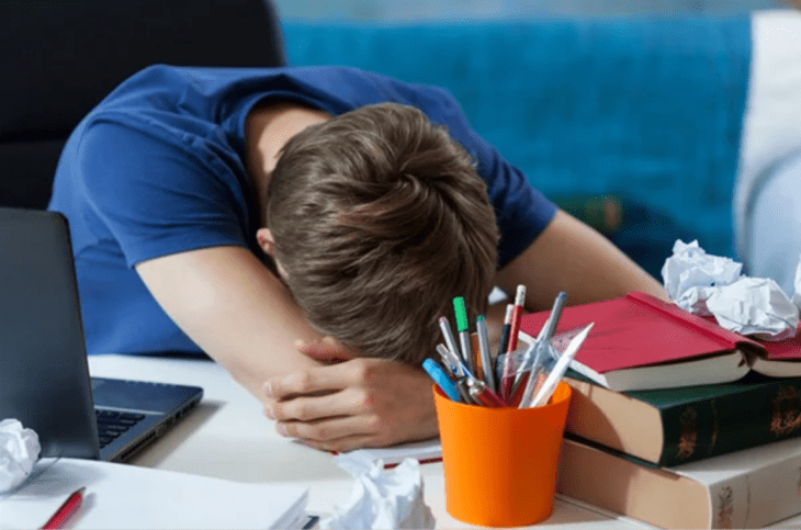 a young student defeated by excessive study leans against his desk to sleep wears a royal blue shirt