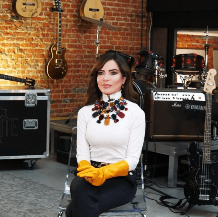 the singer Gloria Trevi sitting in a studio full of musical instruments she is wearing a white blouse and black pants 