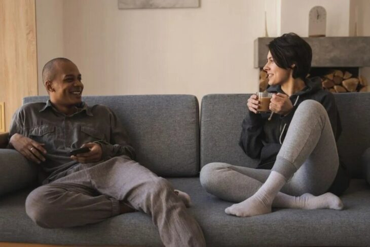 imagen de una pareja sentada en un sofá mientras la chica toma café el hombre sostiene su celular en la mano 