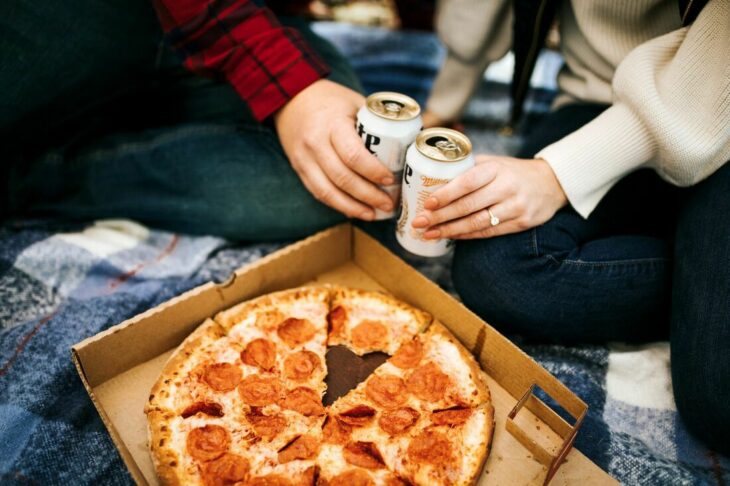 manos de unas personas brindando con un par de latas de cerveza con una pizza 