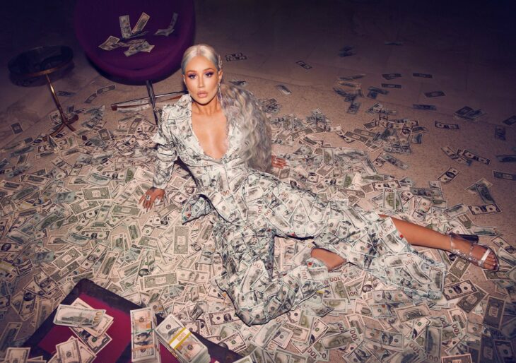 Photograph of Iggy Azalea sitting on the floor surrounded by lots of dollars wearing a 100 dollar bill dress 