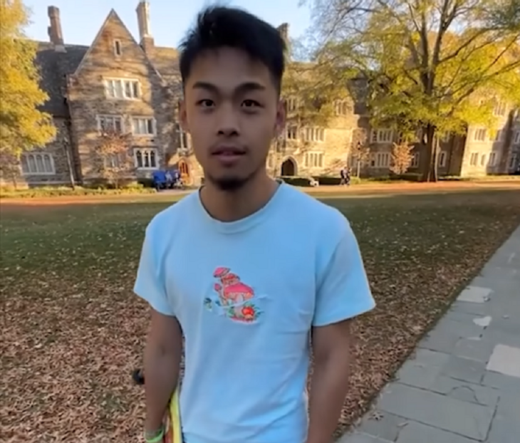 un chico parado en un parque de Estados Unidos lleva una playera azul con un pequeño dibujo es de ascendencia asiática