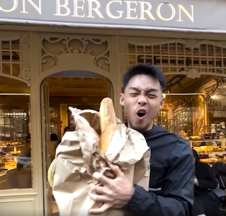 un chico sostiene una bolsa con baguettes afuera de una panadería parisina el chico lleva una sudadera negra y está gritando de alegría