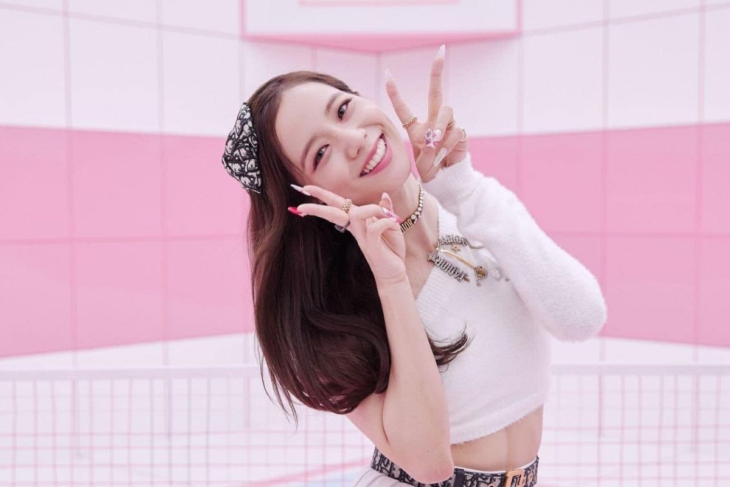 BLACKPINK's Jisoo in an image where she is waving with both hands making the v for victory, she is wearing a white blouse and a black and white bow on her head behind her is a wall painted in pink