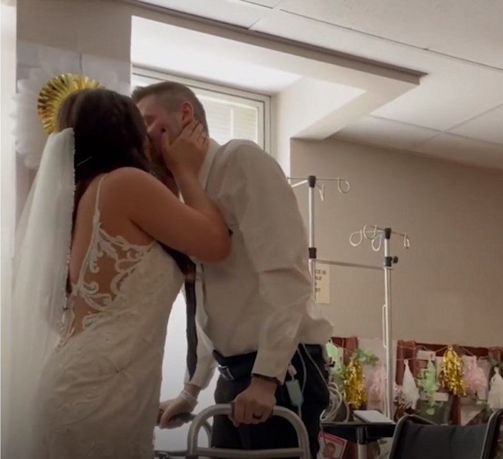 A couple kiss in a hospital room where they got married. He carries a walker to support himself. She is wearing her white wedding dress with a veil. The young man is wearing black pants and a white shirt with a dark tie.