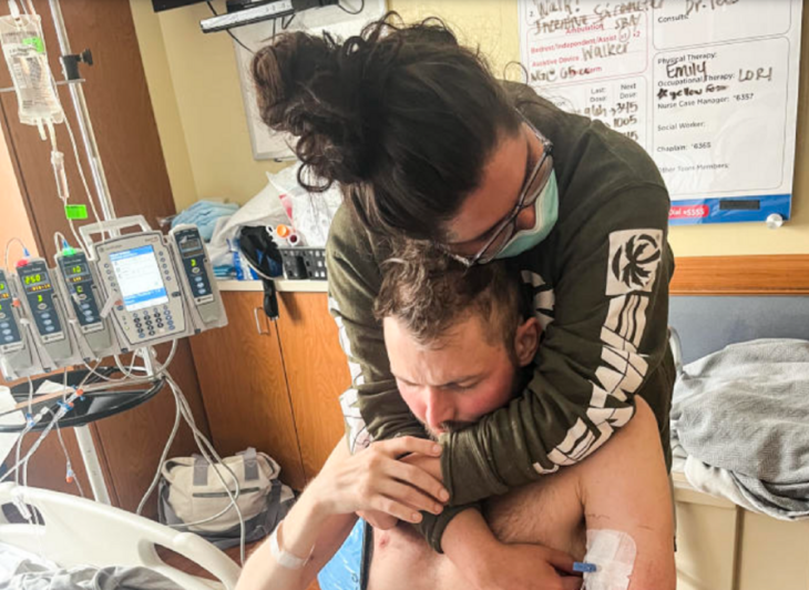 a couple embraces affectionately in a hospital she is wearing an olive green sweatshirt and her hair is tied up in a bun she is wearing a blue face mask and transparent glasses he is bare-chested and has some wires connected to his body