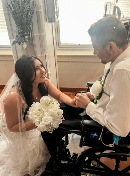 unos novios se están casando en una habitación de hospital ella lleva un vestido largo de novia blanco y velo largo trae un ramo de flores blancas el lleva un pantalón negro y camisa blanca con corbata oscura lleva unas flores blancas en el pecho ella está en cuclillas para estar a la altura del novio