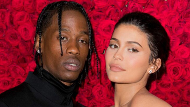 In an image of their faces together, Kylie Jenner and Travis Scott behind them are countless red roses, he is wearing a black suit and both have their hair up, her makeup is natural