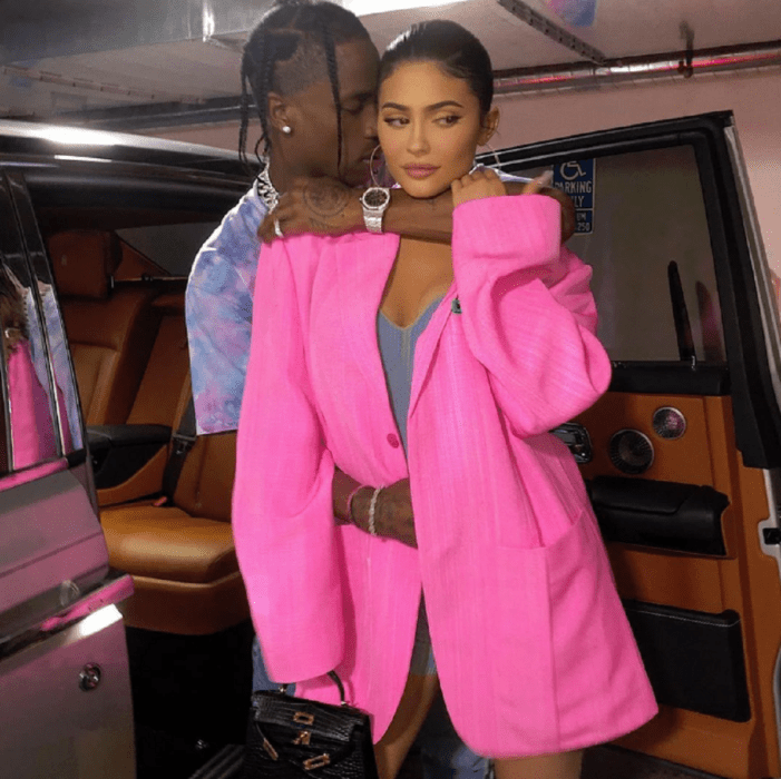 Kylie Jenner and Travis Scott in an image where he is taking her by the neck and affectionately approaches her face she is wearing a long fuchsia jacket he is wearing a patterned shirt they both have their hair up