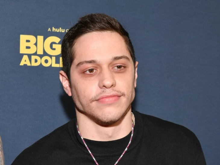 Pete Davidson on a red carpet is dressed in dark clothes and wears a necklace around his neck