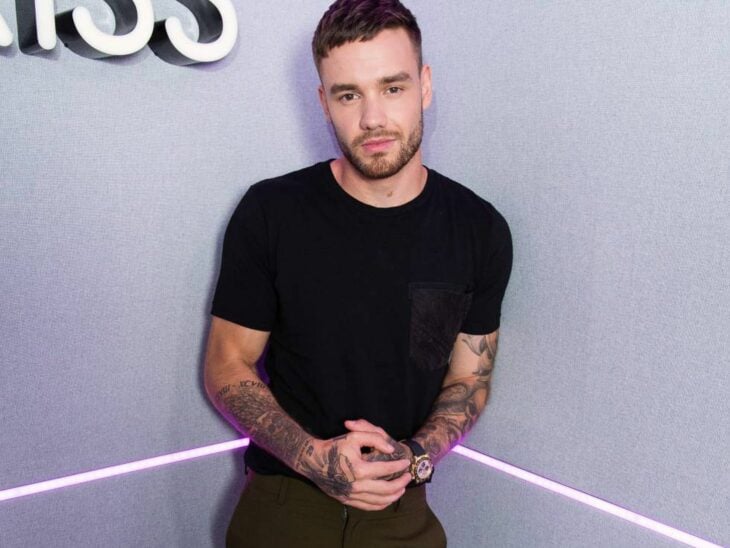 Liam Payne fotografiado en la esquina de una habitación lleva ropa oscura y luce tatuajes en los brazos