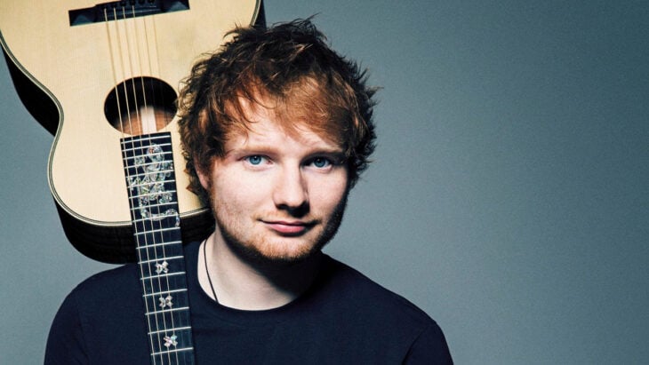 Ed Sheeran holds a guitar upside down with his shoulder wears dark clothing