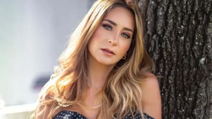 Geraldine Bazán en una imagen donde luce recargada en un árbol lleva el cabello suelto y largo peinado en semi ondas su maquillaje es discreto pero marcado lleva un vestido gris con negro sin mangas ni cuello 