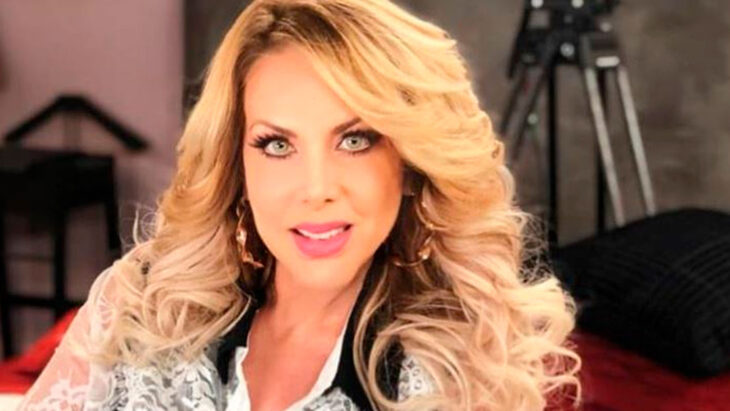 Lorena Herrera en un primer plano lleva un maquillaje muy marcado y el cabello rubio suelto y largo peinado en ondas trae una arracadas grandes doradas está en un estudio con cámaras