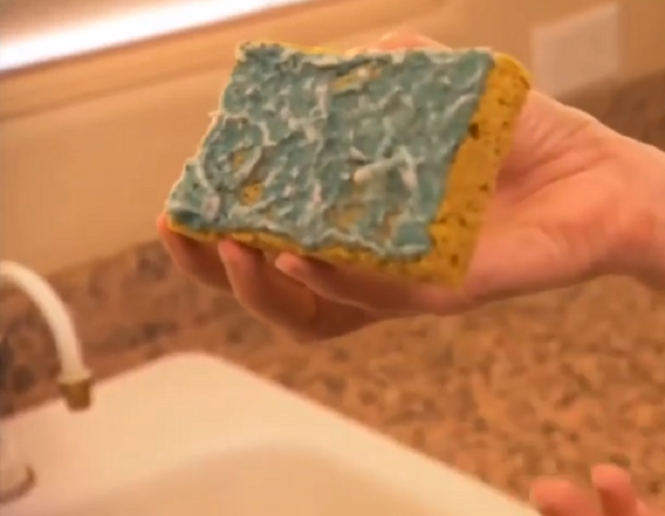 imagen de una gastada esponja para lavar trastes de colores amarillo y verde una mano femenina la sostiene sobre la tarja de la cocina